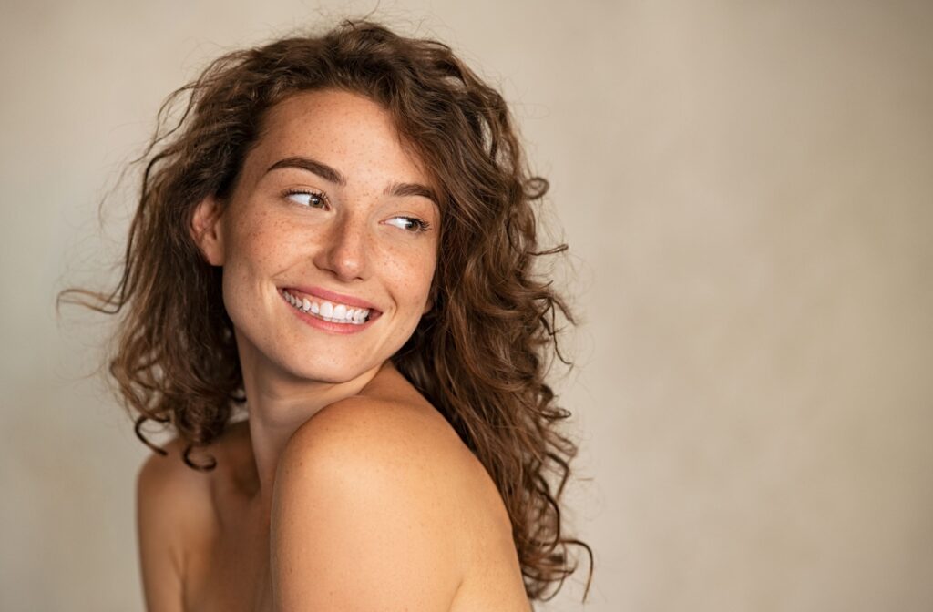 Young person with glowing skin and curly brown hair is smiling wide while looking over their shoulder.