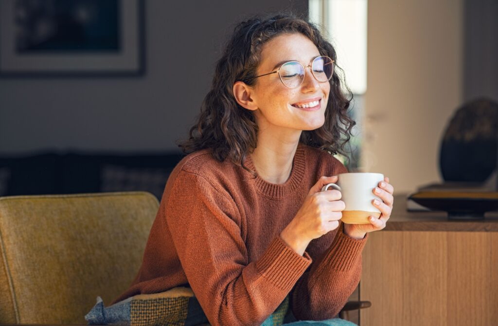 A smiling adult sits on a cozy couch while enjoying a cup of coffee.