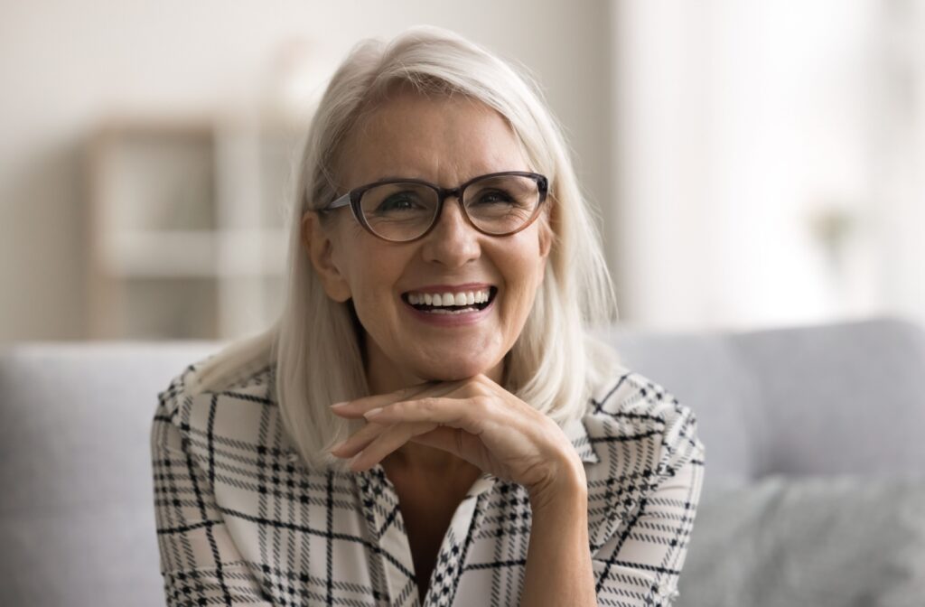 A woman with dentures sitting on a couch, smiling with her hand on her chin.
