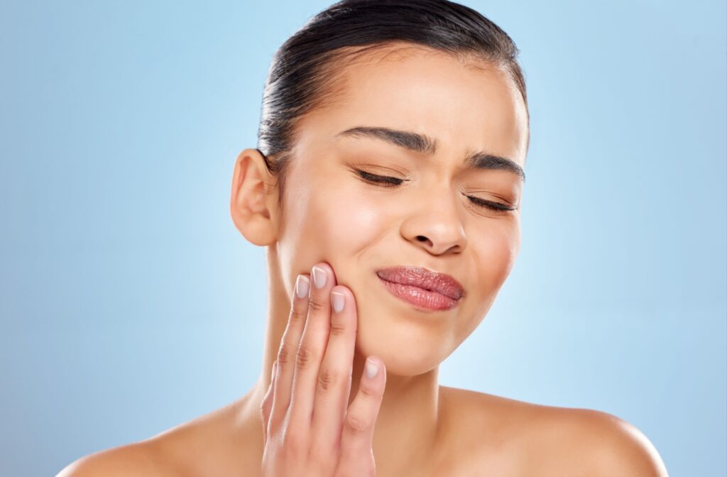 Woman with a toothache touching the side of her face against a blue background.